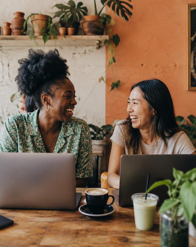 Two friends laughing over coffee while working on laptops in a plant-filled café